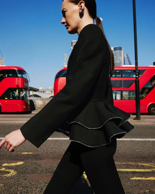 Adult female model in a black suit walks on a crosswalk with red double-decker buses and city buildings in the background on a sunny day.