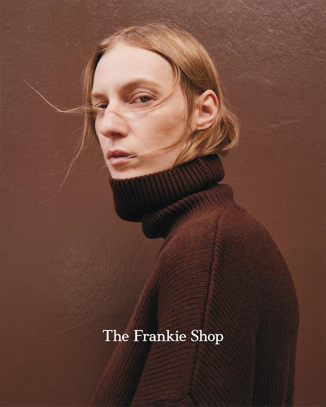 Close-up portrait of a fair-skinned adult in a brown turtleneck sweater against a matching brown background, showcasing fashion editorial style.