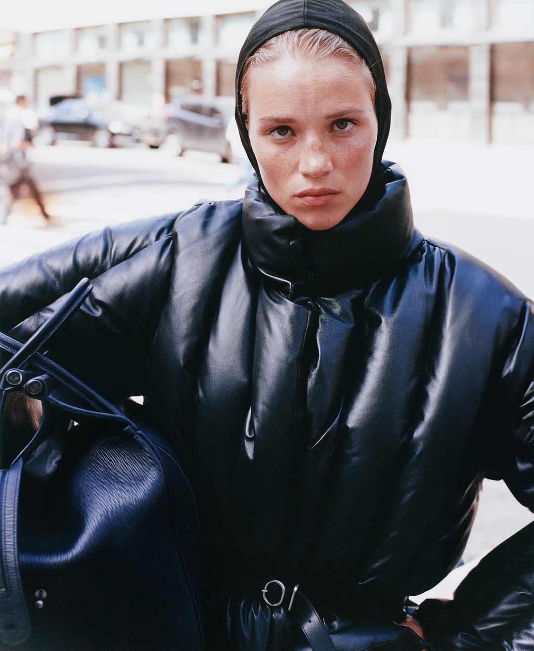 Stylish woman in puffer jacket and headscarf stands on city street holding black leather bag, showcasing urban fashion.
