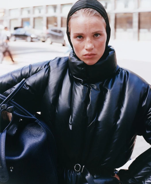 Stylish woman in puffer jacket and headscarf stands on city street holding black leather bag, showcasing urban fashion.