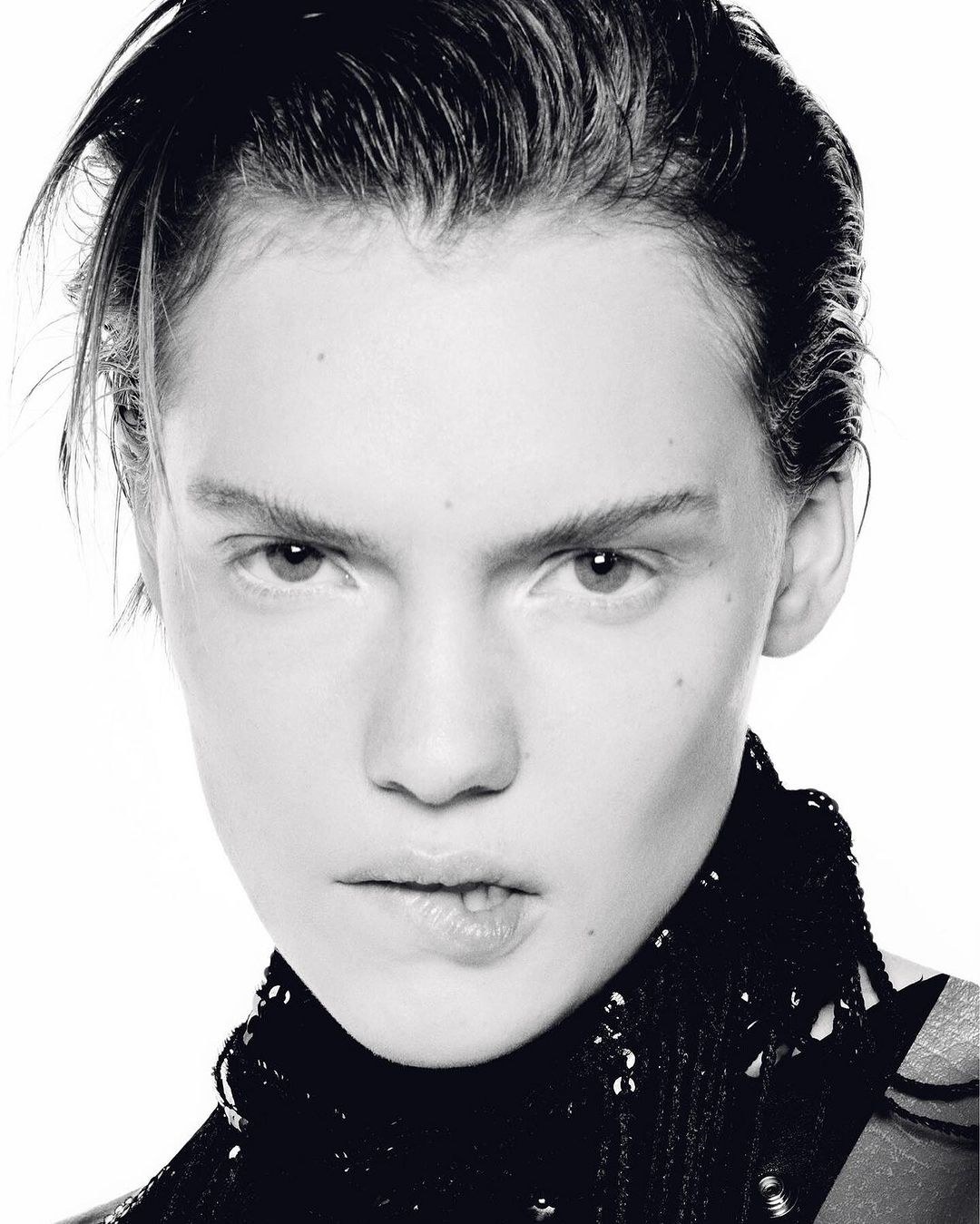 Close-up black and white portrait of a young adult woman with short dark hair, wearing a dark, sequined top against a white background.