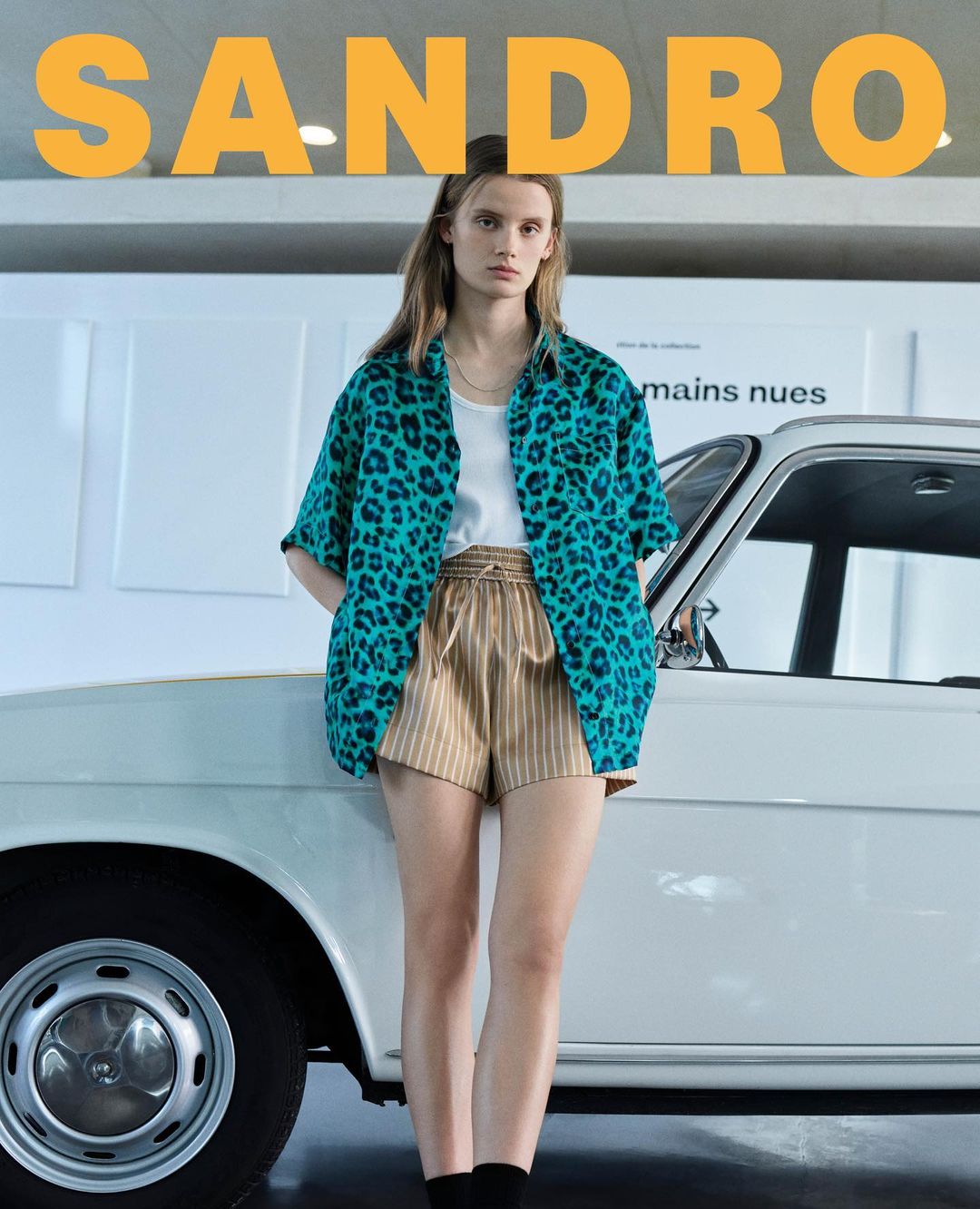 Fashion model wears a turquoise animal print shirt and striped shorts, posing next to a vintage white car in a studio setting.