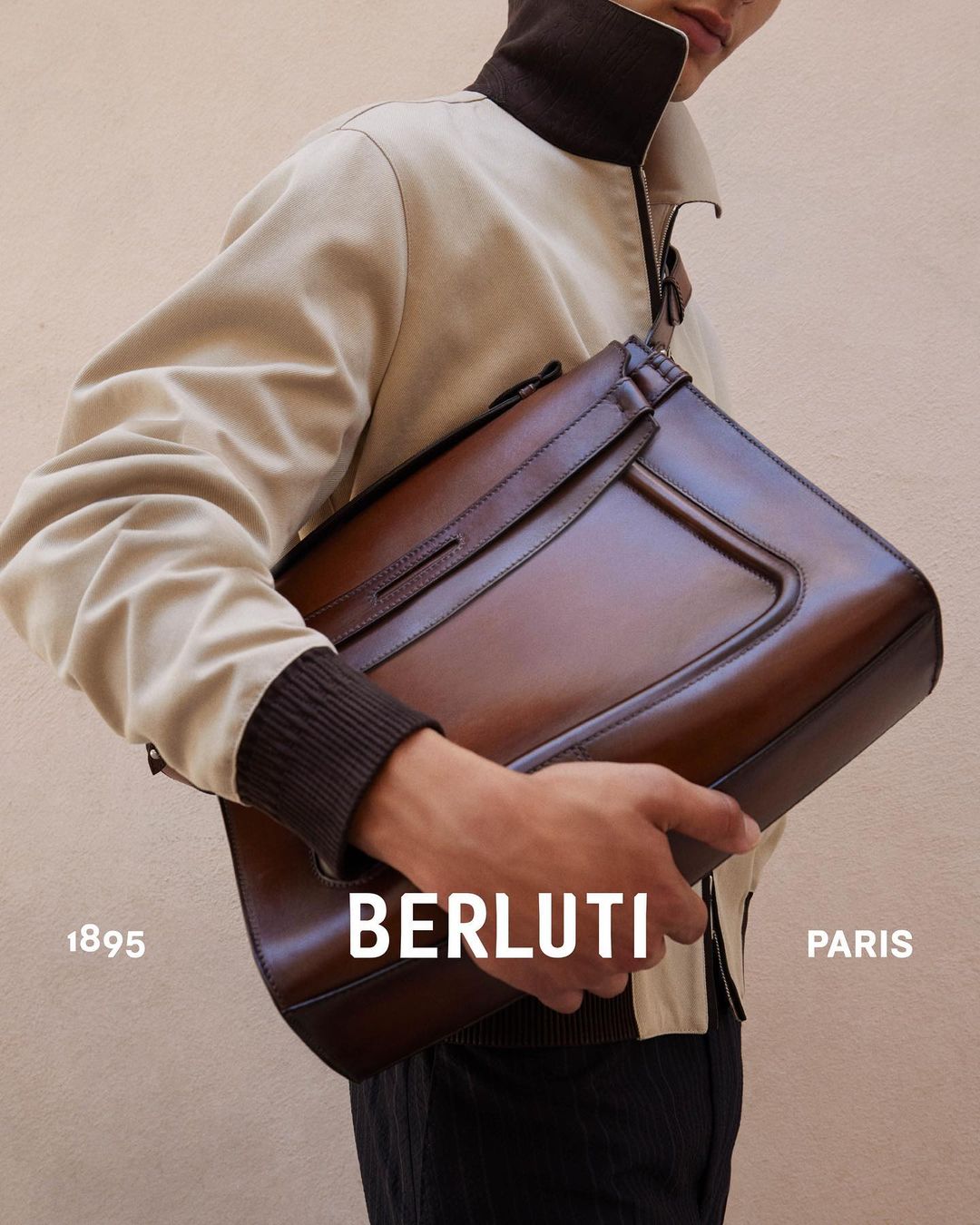 Close-up studio shot of a man holding a brown leather briefcase in front of a tan wall, showcasing fashion and accessory design.