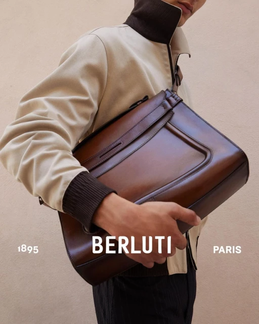 Close-up studio shot of a man holding a brown leather briefcase in front of a tan wall, showcasing fashion and accessory design.