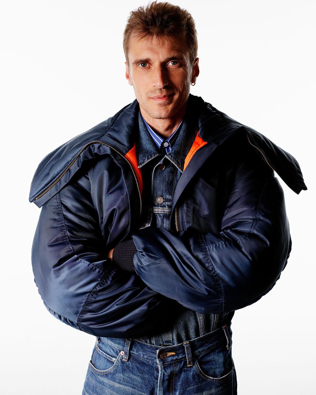 Man wearing a padded navy blue bomber jacket and denim jeans with arms crossed against a plain white background