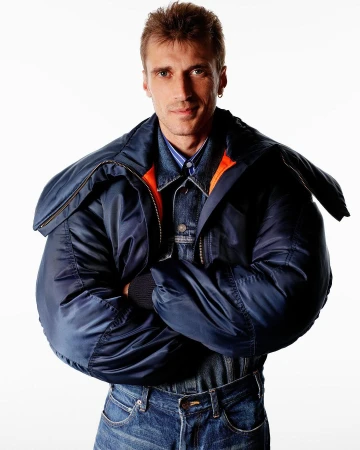 Man wearing a padded navy blue bomber jacket and denim jeans with arms crossed against a plain white background