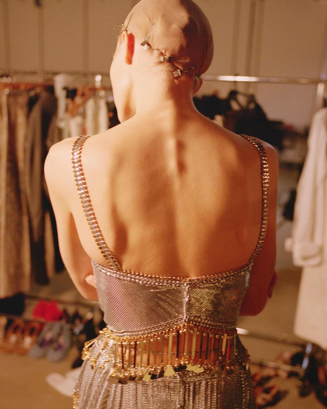 Back view of a slender adult woman wearing a silver and gold jeweled gown and a nude-colored wig cap in a backstage fashion setting.