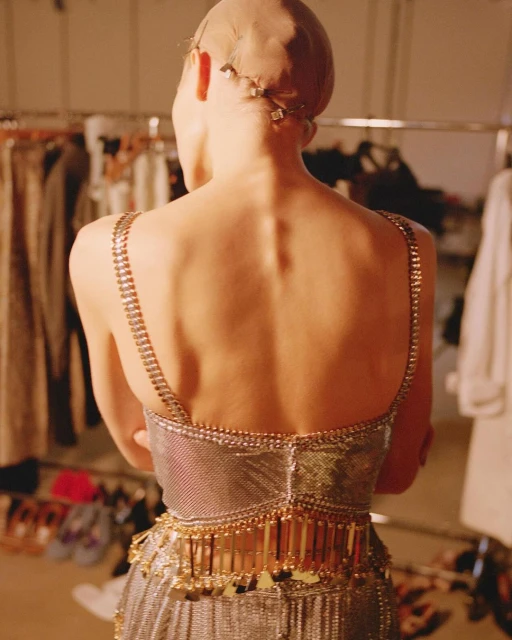 Back view of a slender adult woman wearing a silver and gold jeweled gown and a nude-colored wig cap in a backstage fashion setting.
