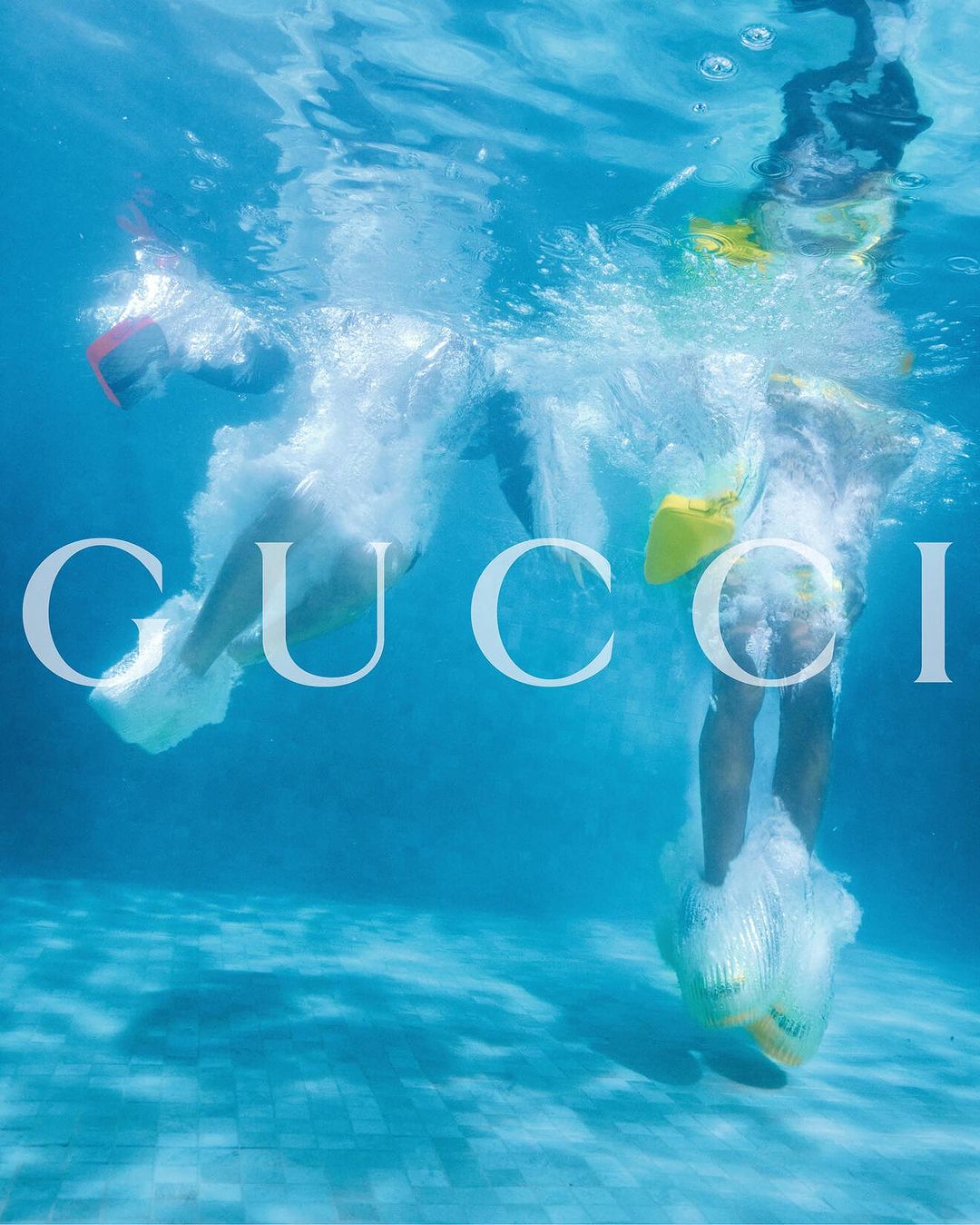 Underwater shot of two people in a pool with Gucci logo overlayed. One person with yellow flippers diving into a pool.