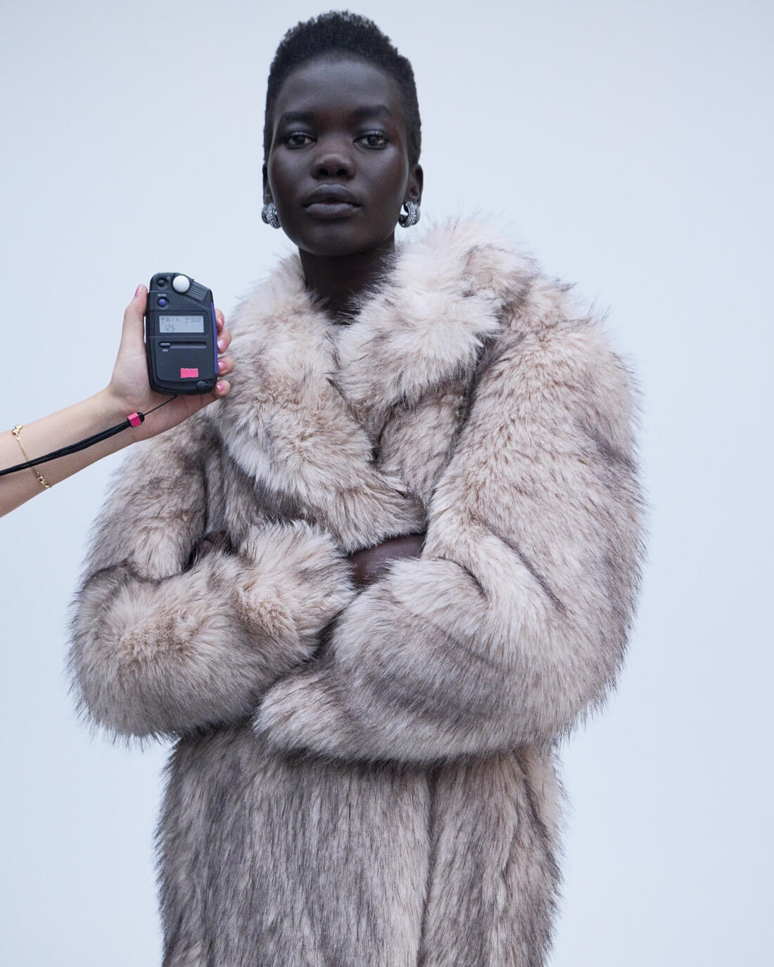 A dark-skinned woman in a furry gray coat poses in front of a gray wall while a light meter is held in front of her face.