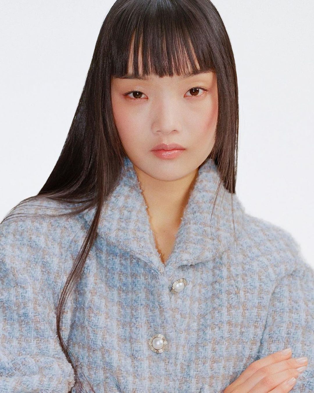 Close-up portrait of an Asian woman with bangs wearing a patterned blue jacket with pearl buttons against a plain white background.