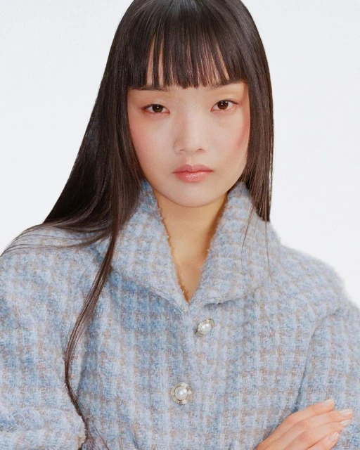 Close-up portrait of an Asian woman with bangs wearing a patterned blue jacket with pearl buttons against a plain white background.