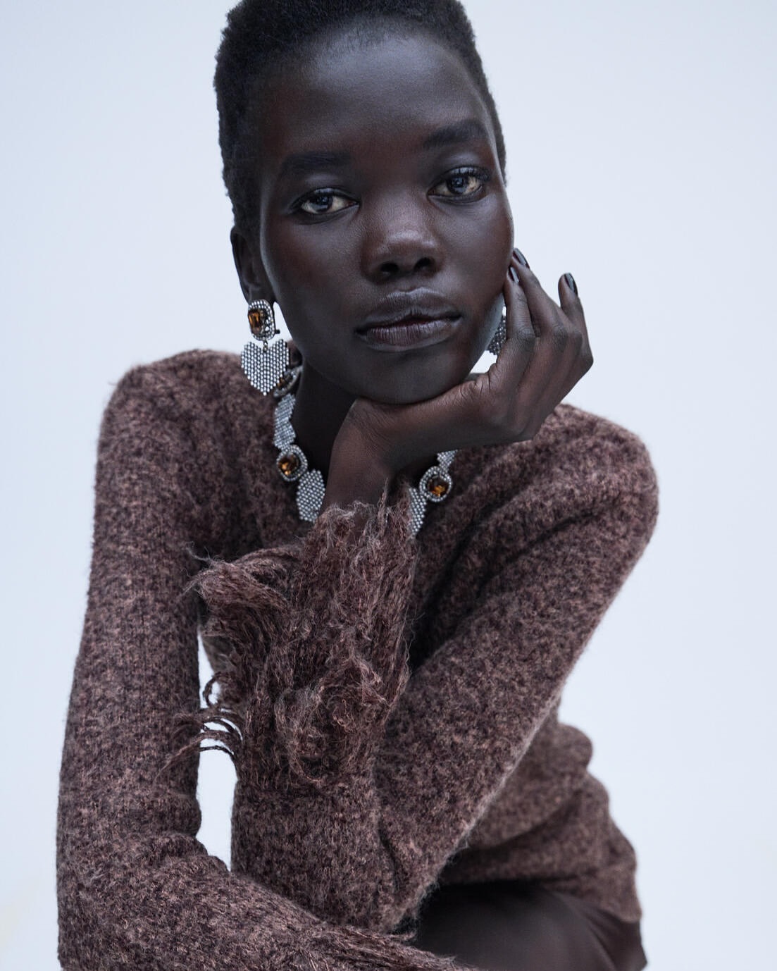 Close-up portrait of a black female model with dark skin wearing brown knit sweater and statement jewelry against white backdrop.
