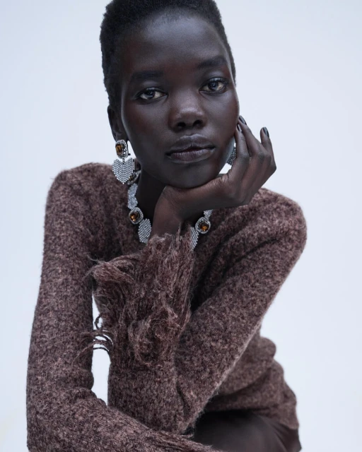 Close-up portrait of a black female model with dark skin wearing brown knit sweater and statement jewelry against white backdrop.