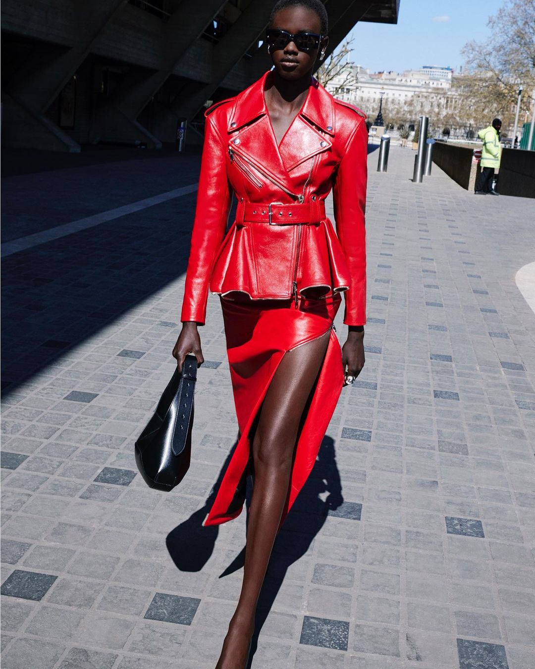 An adult woman poses outside wearing a red leather jacket and skirt, accessorized with sunglasses and a black handbag on a sunny day.