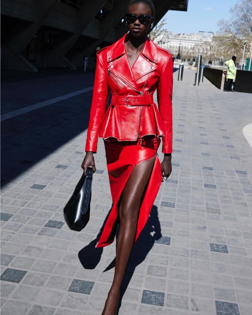 An adult woman poses outside wearing a red leather jacket and skirt, accessorized with sunglasses and a black handbag on a sunny day.