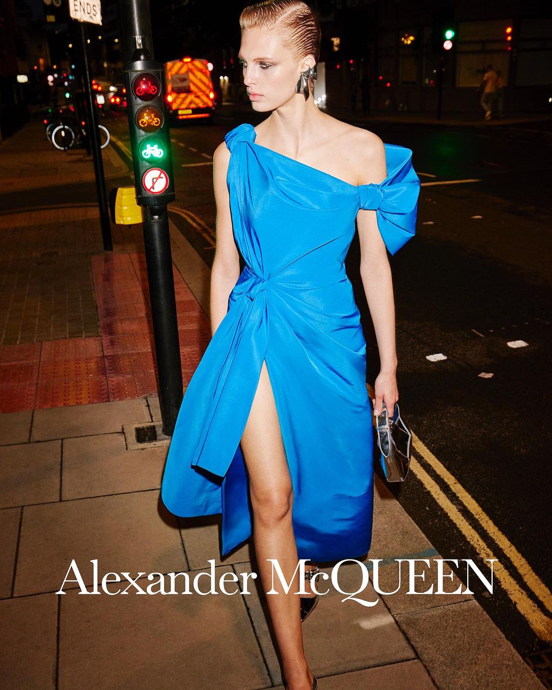 A blonde fashion model poses on a city street at night wearing a bright blue Alexander McQueen dress with traffic lights and an ambulance in the background.