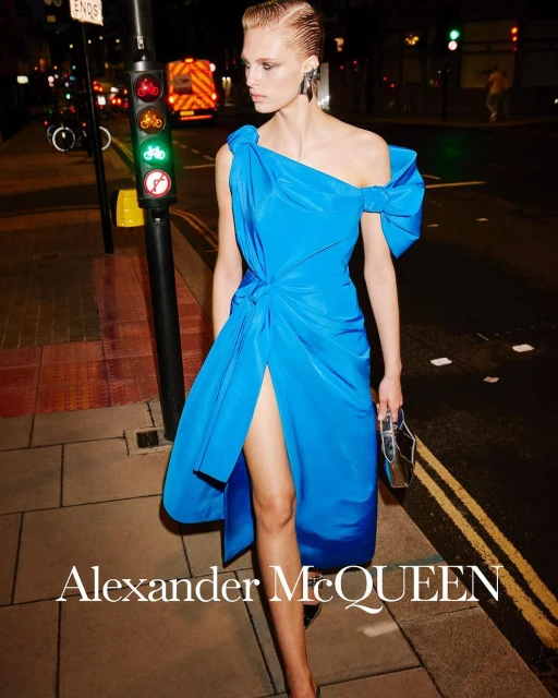 A blonde fashion model poses on a city street at night wearing a bright blue Alexander McQueen dress with traffic lights and an ambulance in the background.