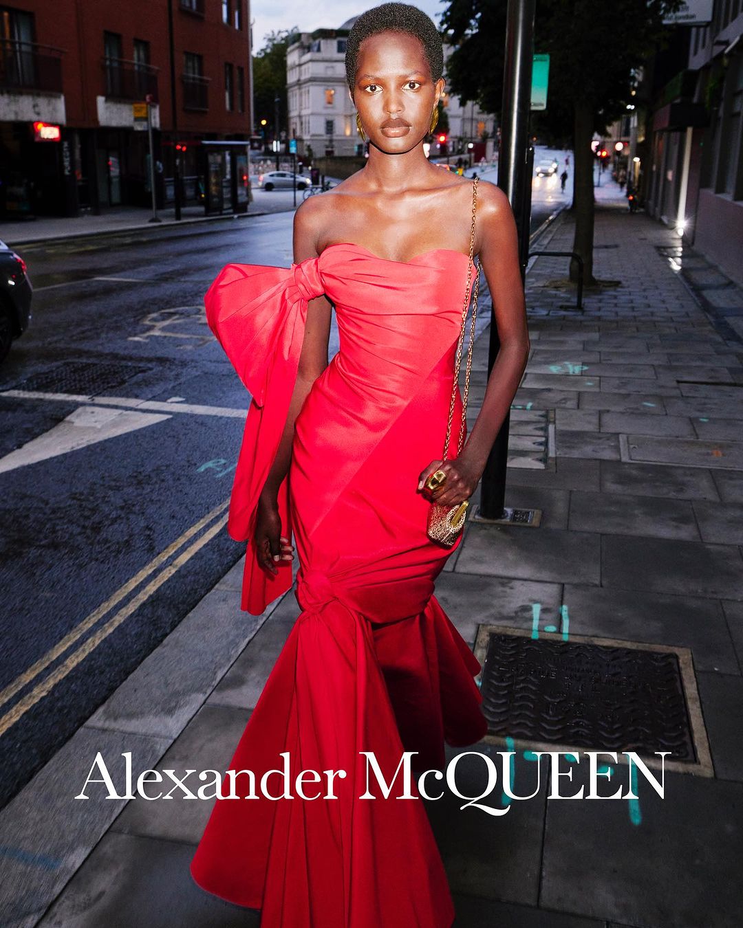 Elegant woman in red strapless gown with bow poses on city street in evening light. Accessorized with gold jewelry, small purse.