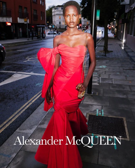 Elegant woman in red strapless gown with bow poses on city street in evening light. Accessorized with gold jewelry, small purse.