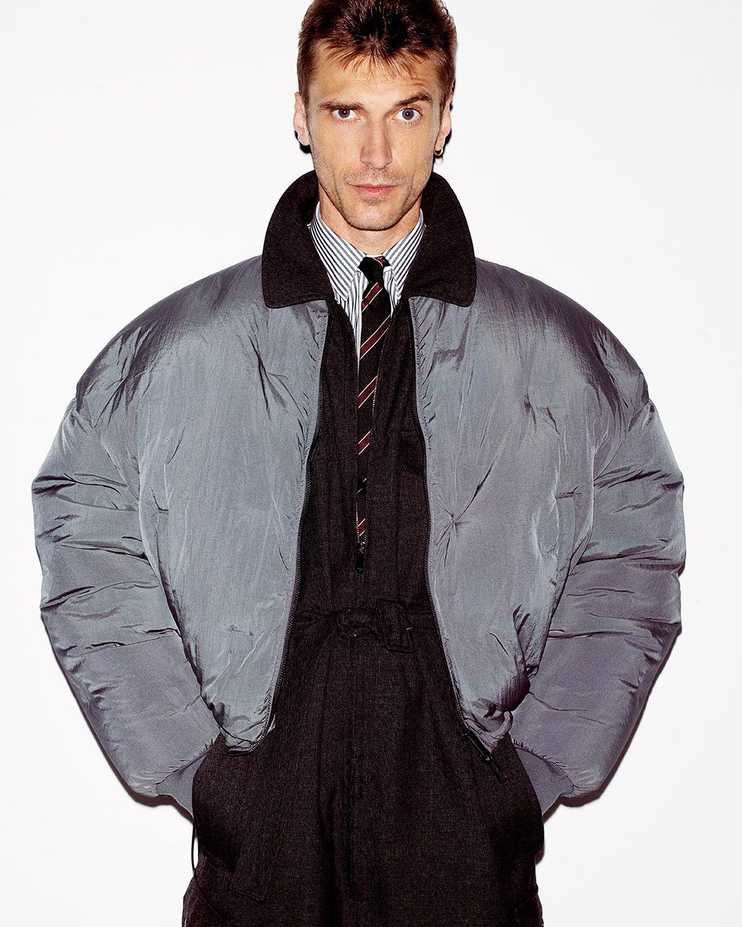 Portrait of a slender adult man in a shiny grey puffer jacket, striped dress shirt, dark tie, and matching trousers against white.