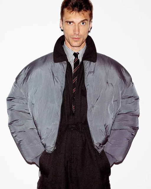 Portrait of a slender adult man in a shiny grey puffer jacket, striped dress shirt, dark tie, and matching trousers against white.