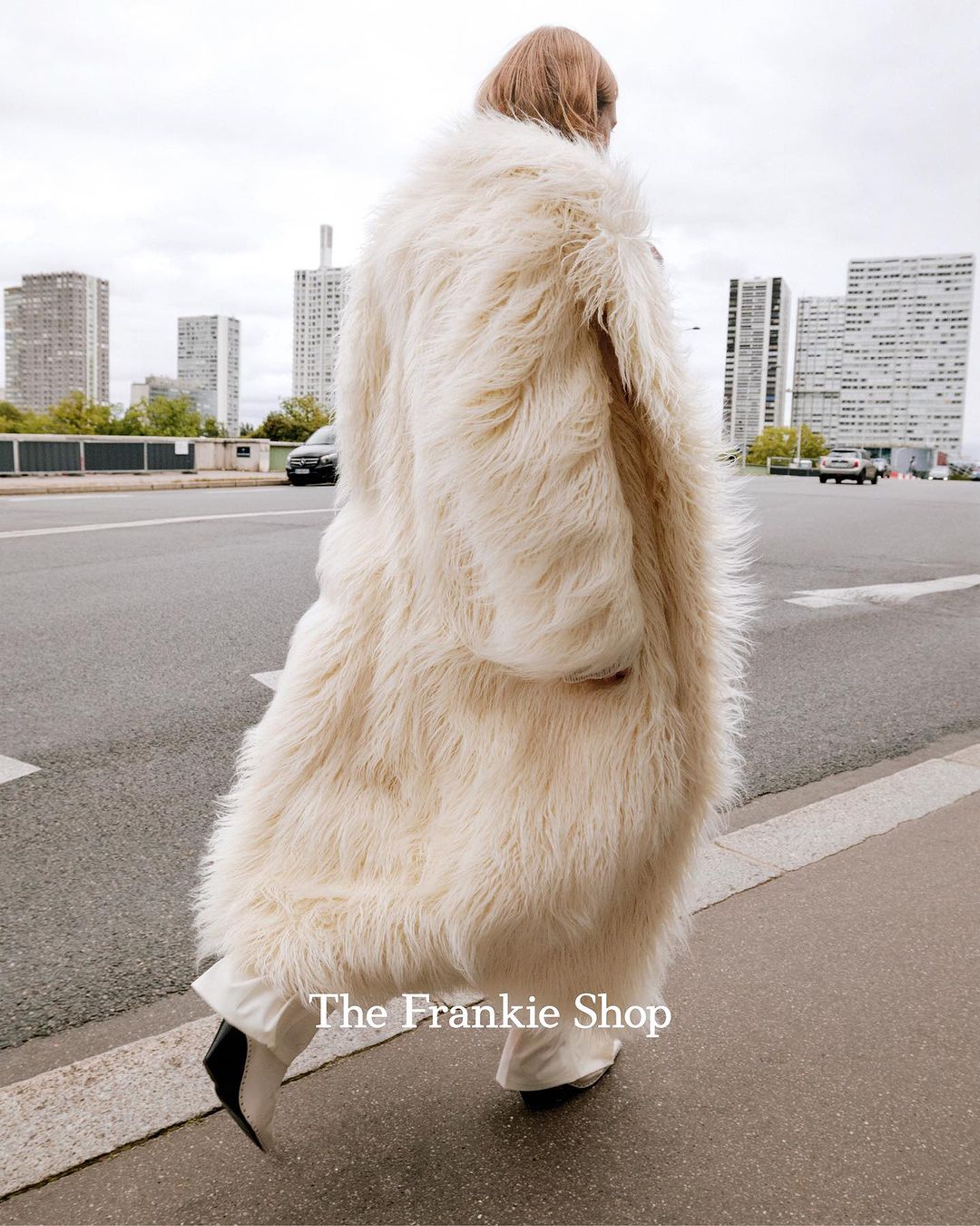 A person wears an oversized, cream-colored, shaggy coat on a city street with tall buildings in the distance on an overcast day.