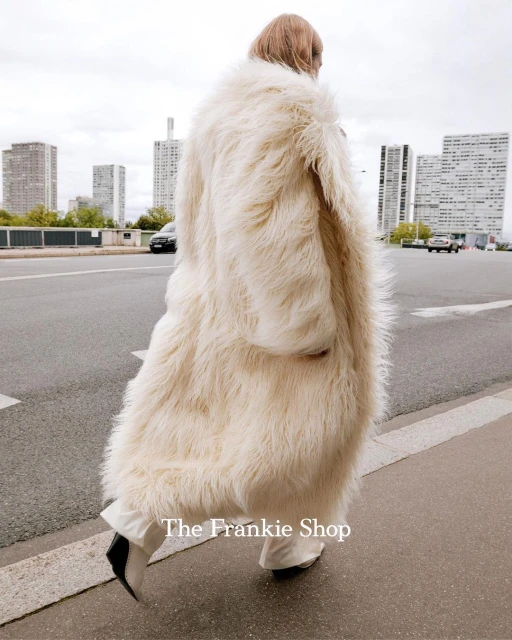 A person wears an oversized, cream-colored, shaggy coat on a city street with tall buildings in the distance on an overcast day.