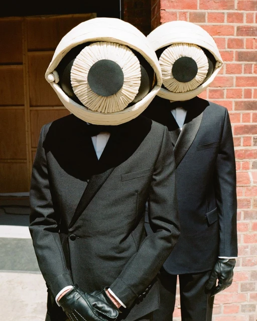 Two figures in black suits and spherical eye masks stand against a brick wall, creating a surreal, formal, and slightly unsettling scene.