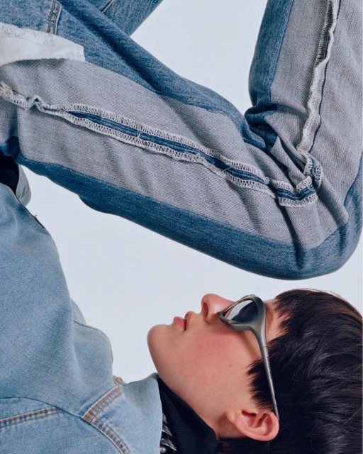 Fashion shot of a model wearing sunglasses, a denim jacket, and patchwork jeans against a white backdrop, showcasing modern apparel.