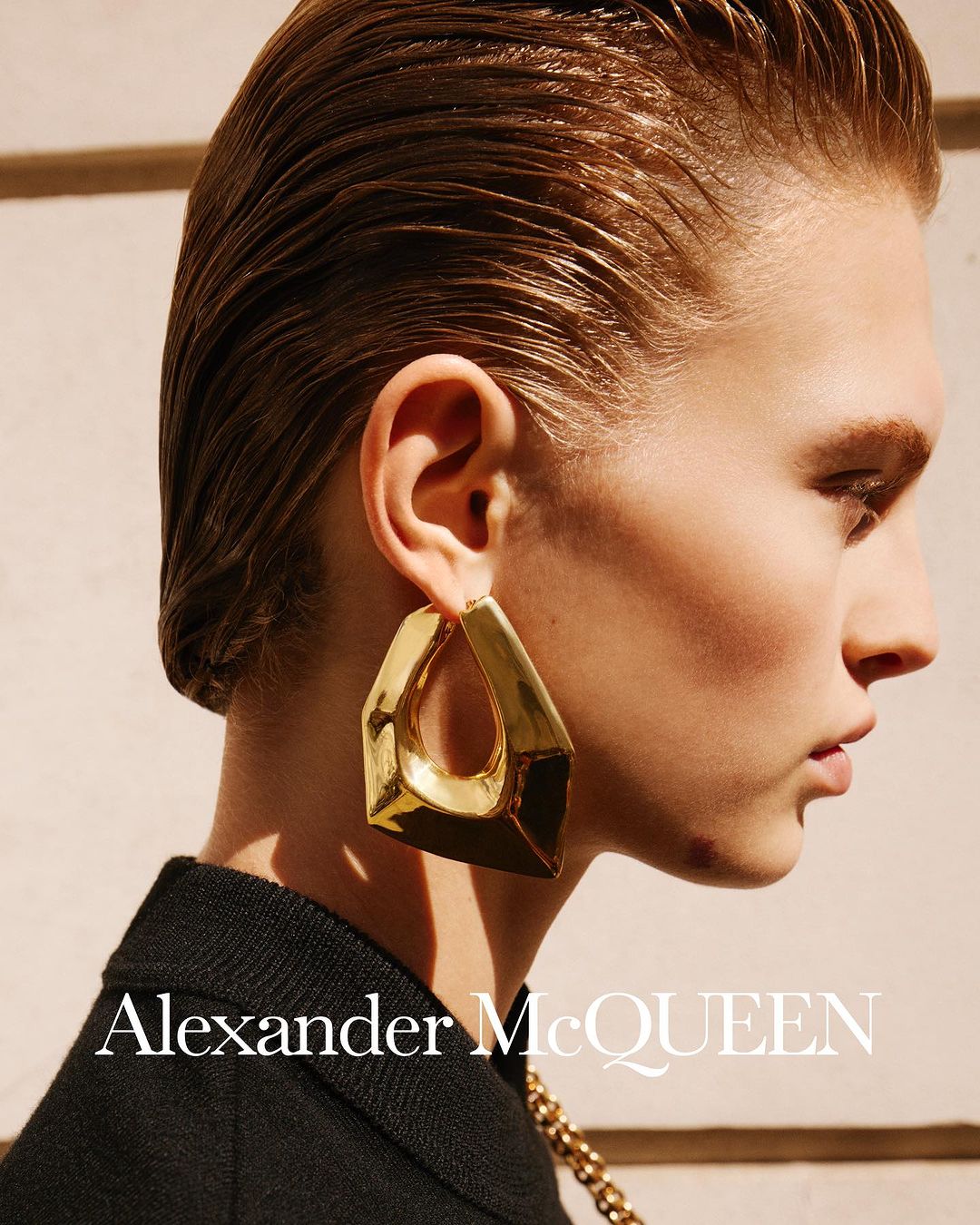 Profile shot of a fair-skinned woman with slicked-back hair and wearing an over-sized gold earring, with the Alexander McQueen logo visible.