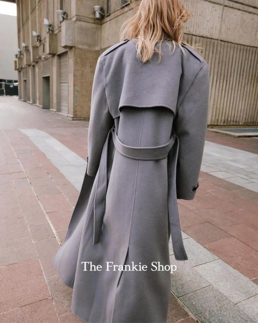 Back view of a blonde woman in a stylish grey belted wool coat walking in an urban setting. Fashion editorial photography.