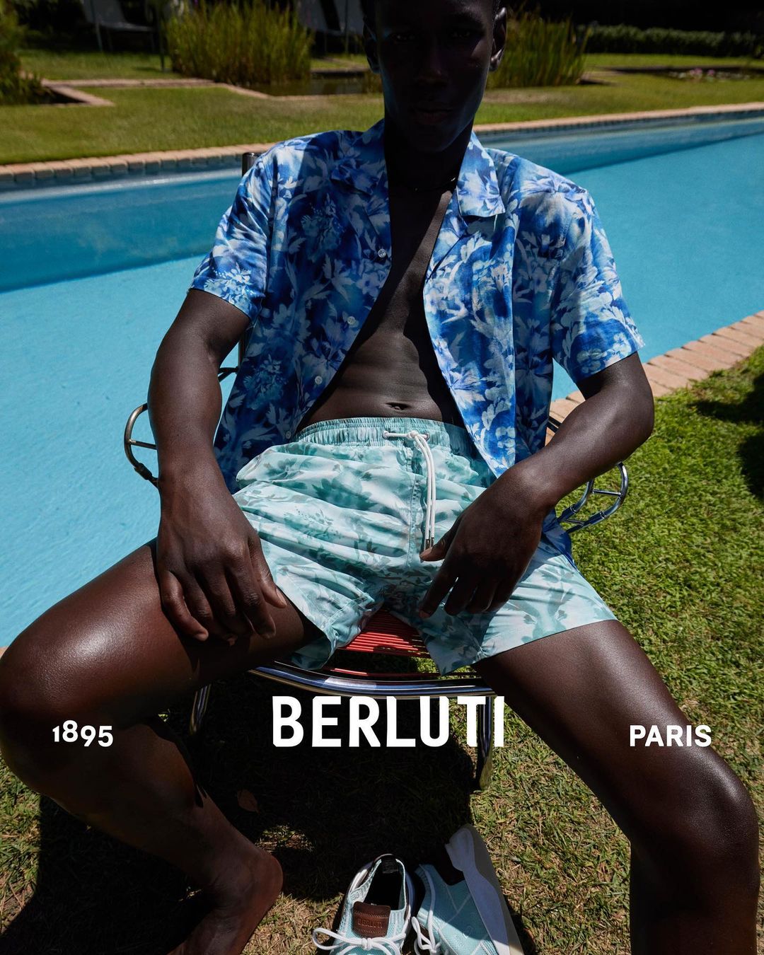A dark-skinned man in a floral shirt and swim trunks sits outdoors near a pool with designer shoes, showcasing high-end fashion.