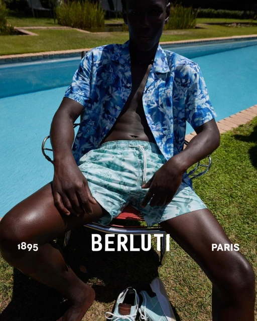 A dark-skinned man in a floral shirt and swim trunks sits outdoors near a pool with designer shoes, showcasing high-end fashion.