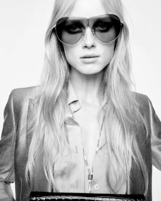 Monochrome portrait of a blonde adult woman wearing sunglasses, a blazer, and a necklace, holding a black clutch against a white background.