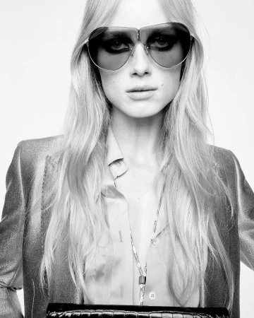 Monochrome portrait of a blonde adult woman wearing sunglasses, a blazer, and a necklace, holding a black clutch against a white background.