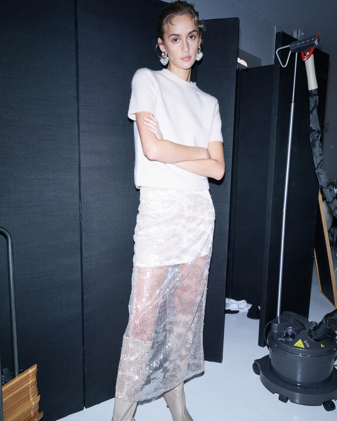 A young woman in a white top and sheer skirt poses in an indoor setting against a dark backdrop. Fashion shot, studio environment.