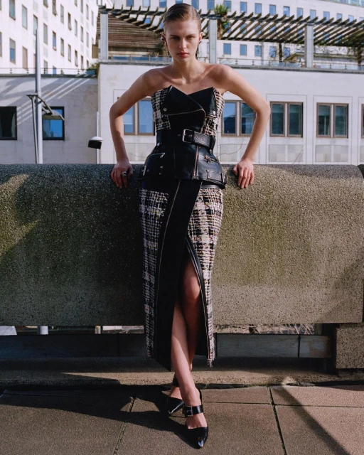 Fashion editorial shot of a blond woman in a strapless midi dress with a leather corset belt, posing on a concrete ledge in the city.
