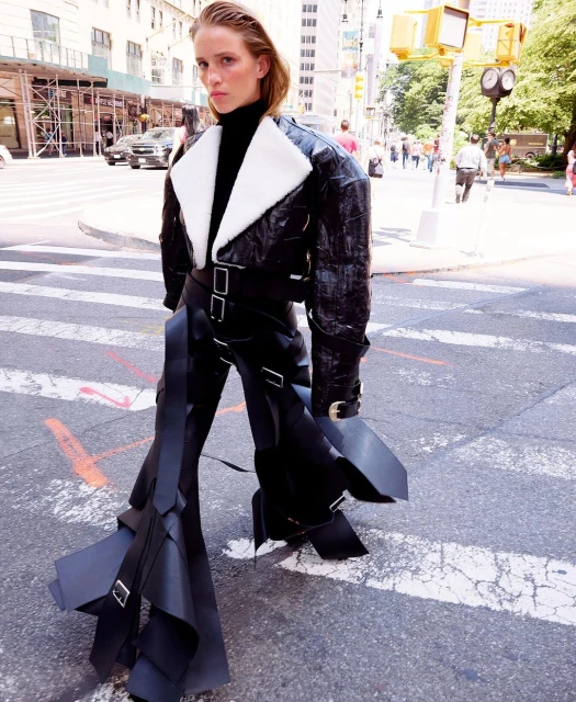 Fashion model in black leather jacket and pants with unusual design posing on a city street with buildings and pedestrian traffic.