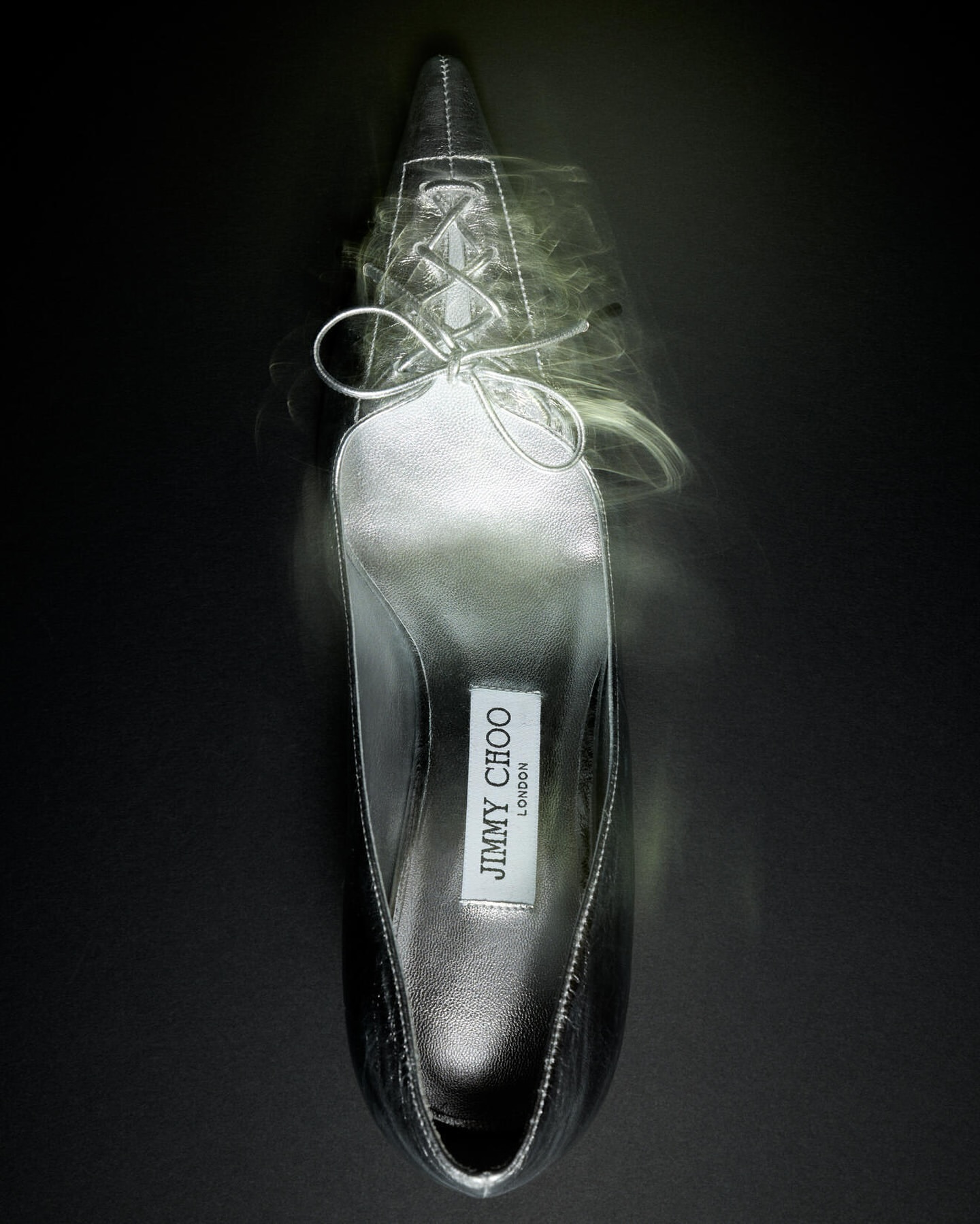 Elevated view of a silver Jimmy Choo stiletto pump against a dark background with a blurred, cloudy effect around the shoe.