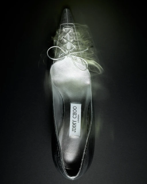 Elevated view of a silver Jimmy Choo stiletto pump against a dark background with a blurred, cloudy effect around the shoe.