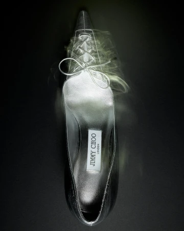 Elevated view of a silver Jimmy Choo stiletto pump against a dark background with a blurred, cloudy effect around the shoe.