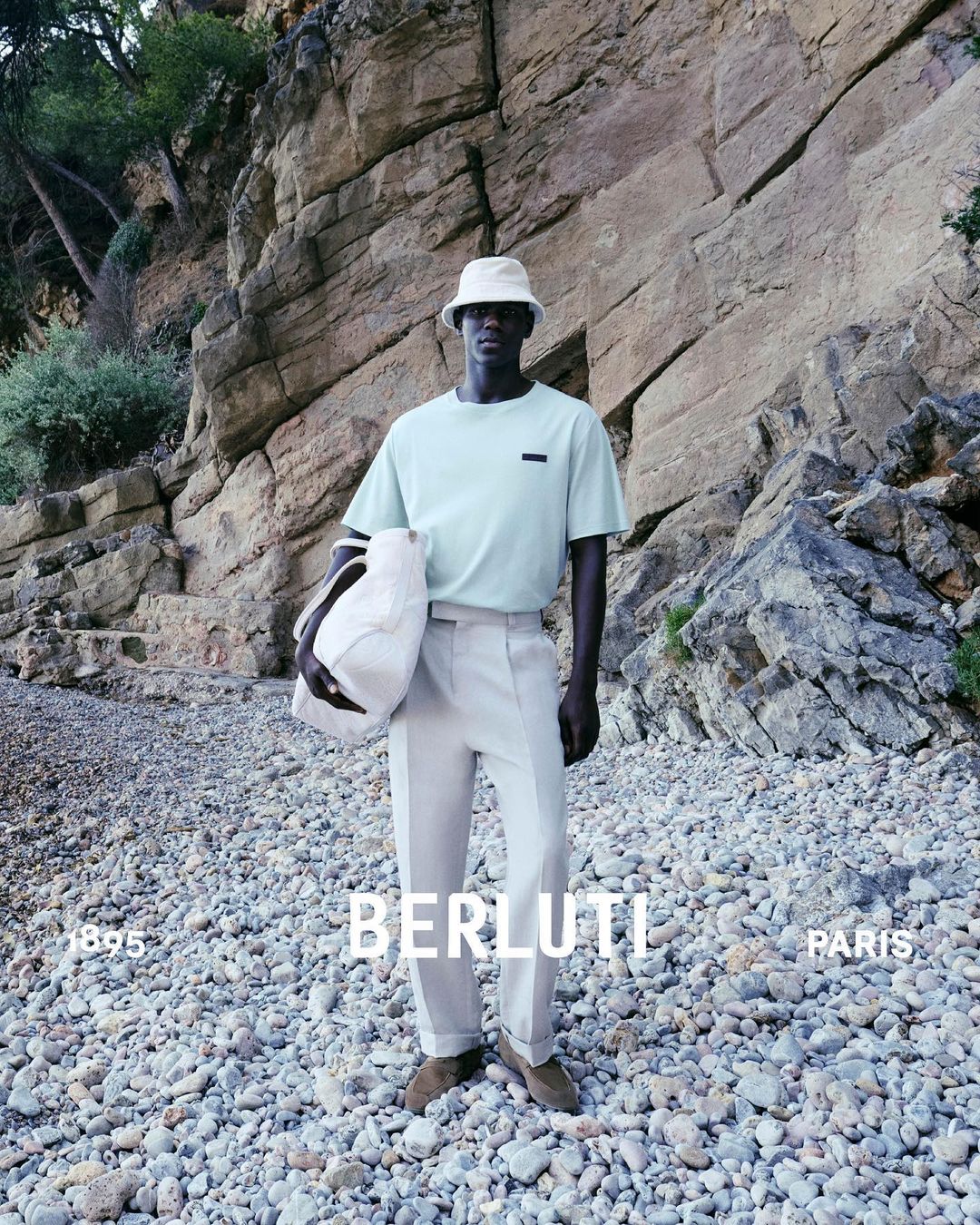 Man in light green shirt and bucket hat stands on rocky beach, holding a tote bag, with rocky cliffs in background for Berluti.