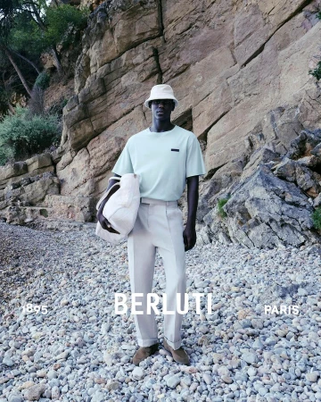 Man in light green shirt and bucket hat stands on rocky beach, holding a tote bag, with rocky cliffs in background for Berluti.