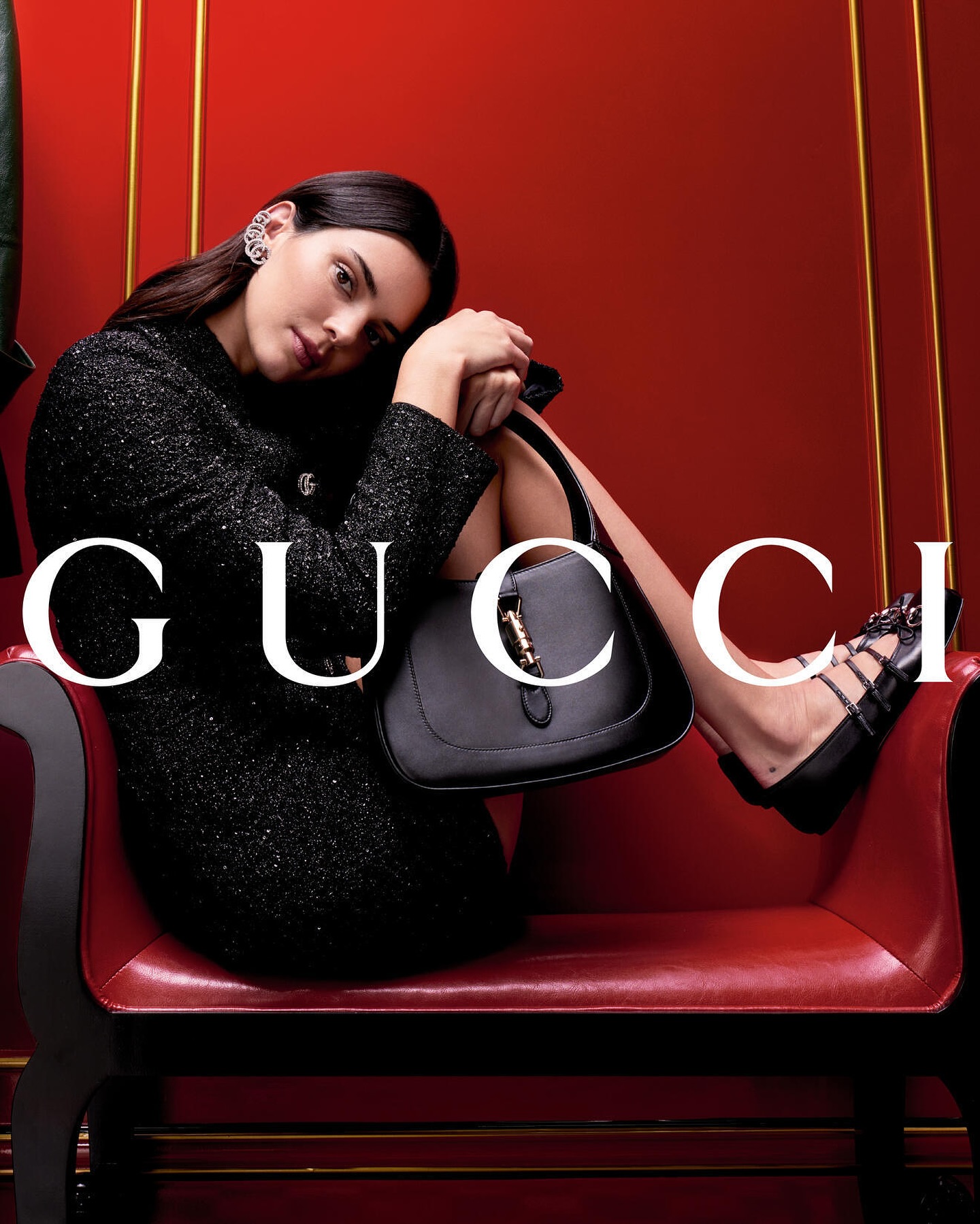 Model Kendall Jenner wearing black textured dress holding black handbag seated on red chair