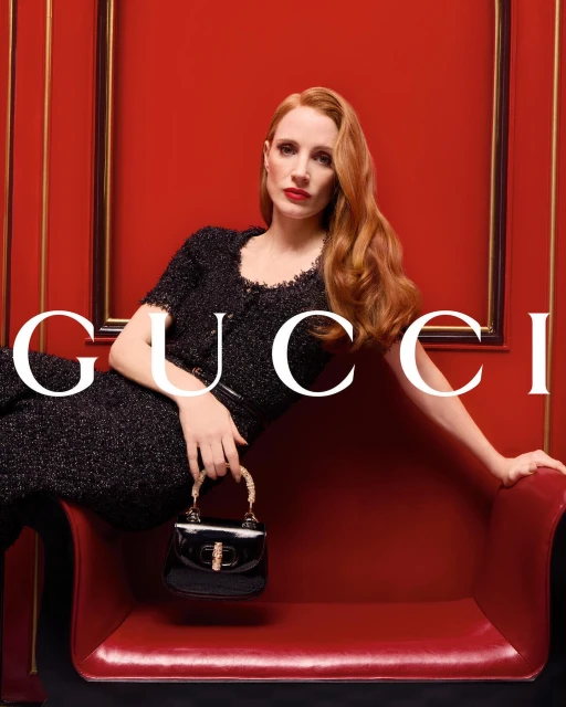 Elegant woman with red hair in black dress poses with a black purse against a red backdrop. Fashion photography.
