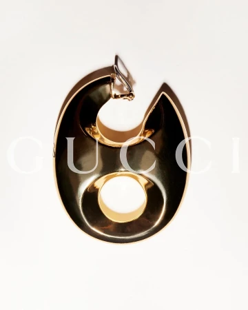 Single, bold gold earring on a white background, featuring a stylized, geometric design with a distinct circular cutout.
