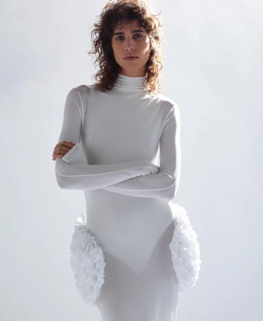 Woman with curly brown hair wearing a long-sleeved white turtleneck dress with ruffled hip accents against a pale background