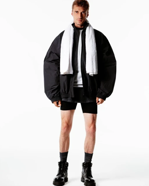 Adult male in a black athletic jacket and black shorts posing indoors with a white towel around his neck, and black boots.