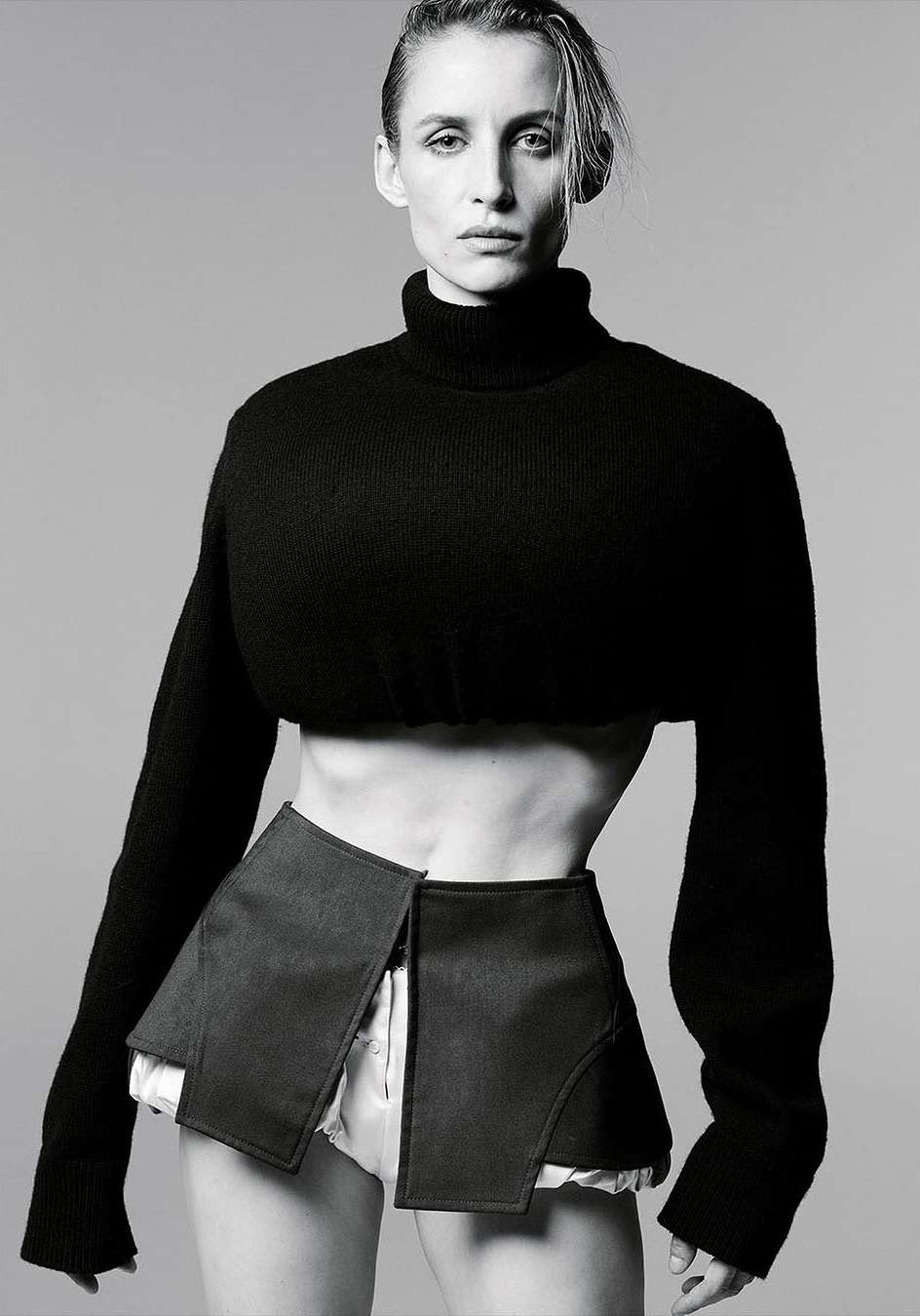 Monochrome fashion portrait of a slim adult woman in a cropped black turtleneck sweater and open black skirt, posing in a studio.